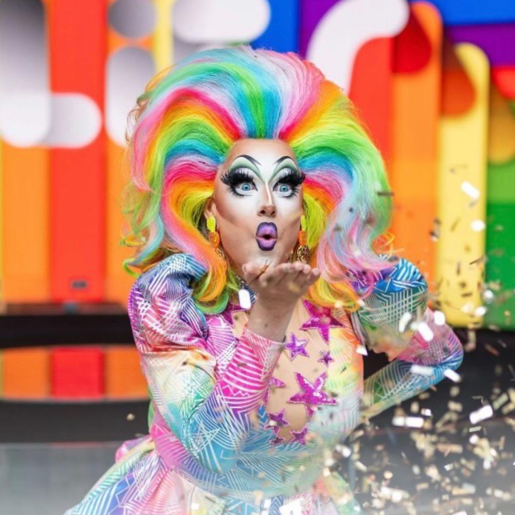 Photo of Kita Mean wearing a rainbow wig and a dress covered in stars. She is blowing glitter through the air.